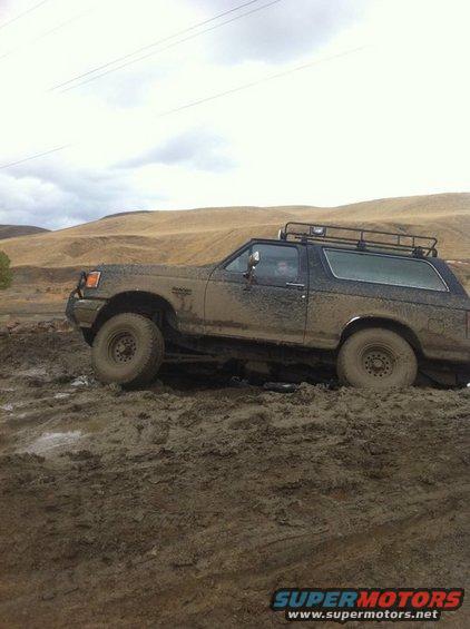 Muddy Ford Bronco with roof rack stuck in a muddy ditch, driver visible inside