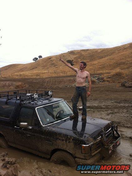 Man standing on the hood of a black 1990 Ford Bronco stuck in deep mud. The vehicle has a roof rack with lights