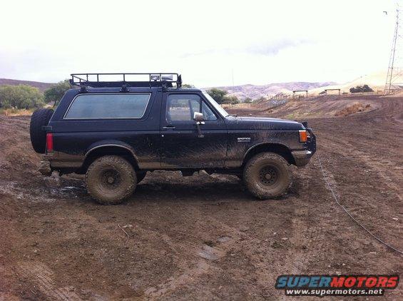 Black 1990 Ford Bronco on muddy terrain with roof rack and spare tire