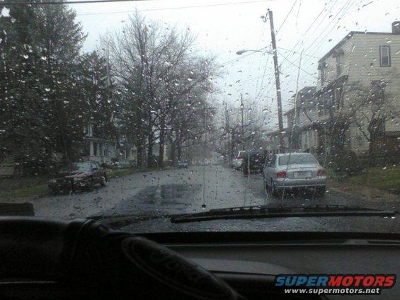 View from inside a car looking out at a rainy street with houses and parked cars
