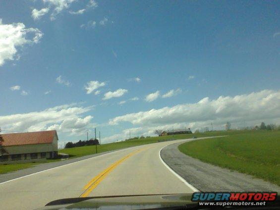 Dashboard view of a road curving through a grassy landscape with a barn under a cloudy sky