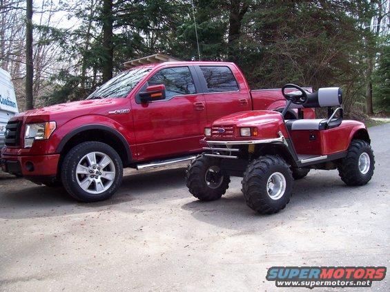 Red pickup truck parked next to a red golf cart with oversized tires. The truck appears to be a Ford F-150 from the 2009-2014 generation