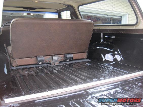 Brown vinyl seat folded up in dark cargo area of an SUV. Ribbed floor, white trim. Watermark 'SUPERMOTORS.NET'