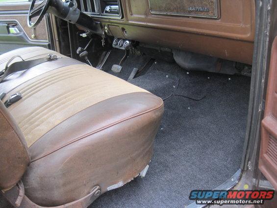 Interior of a vehicle showing the driver's seat, dashboard, pedals, and carpeted floor