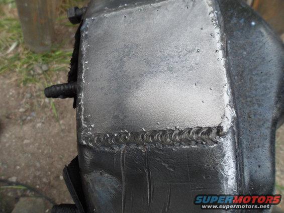 Close-up of a metal part with visible welds and a bolt sticking out. Supermotors.net logo in corner