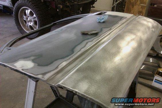 Car door stripped to metal, being prepped for paint. Another vehicle's tire is visible in the background