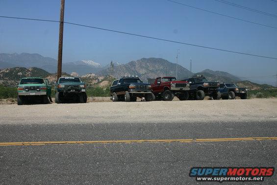 Several lifted trucks parked in a row with mountains behind them. One is a dark blue Bronco