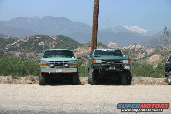 Two green Ford trucks parked on a dirt road, one with "ABUSED" sticker on windshield. Mountains in background