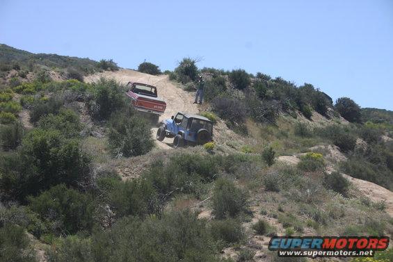 Red pickup truck and blue Jeep climbing a steep dirt trail with brush on either side