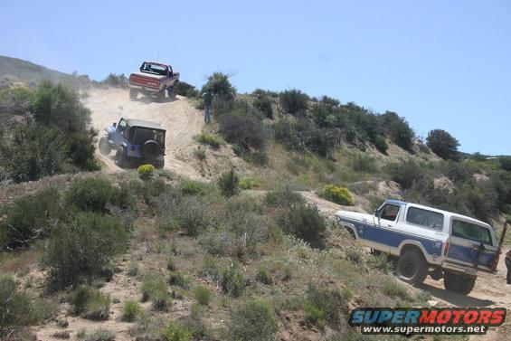 Three off-road vehicles driving up a dusty dirt hill. A blue Jeep, a red pickup truck, and a white and blue Ford Bronco