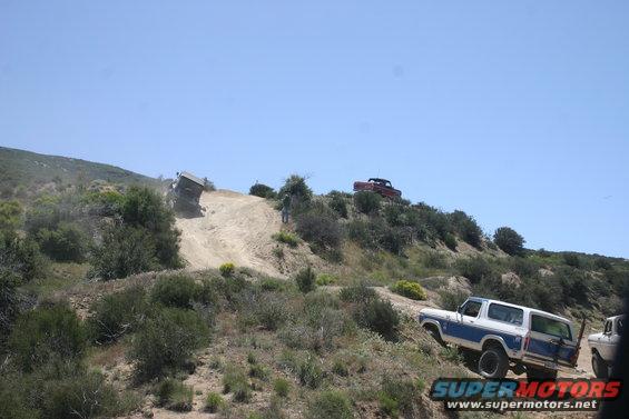 Blue and white SUV climbing a steep dirt hill, another SUV on the crest, and a third vehicle partially visible