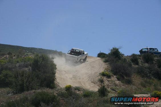 White off-road vehicle kicking up dust while descending a dirt hill, roll cage visible. Another blue and white SUV is parked on the ridge