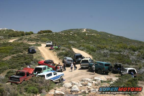 Group of off-road vehicles parked on a dirt road in a hilly, scrub-covered landscape
