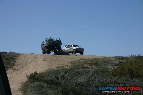 1991 Ford Bronco and a white SUV on a dirt hill under a clear blue sky