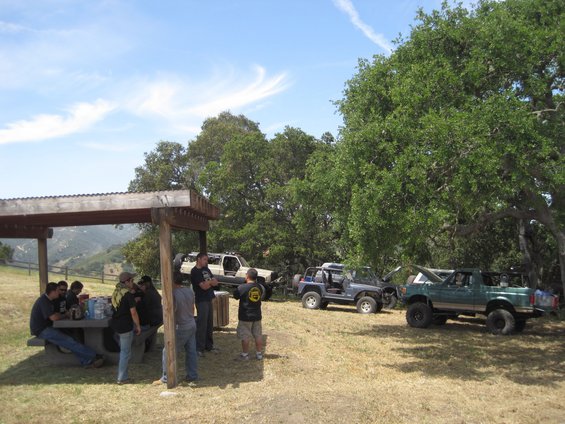People gathered at a picnic table under a shelter, with several off-road vehicles parked nearby under trees