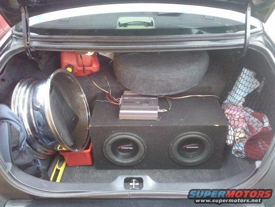 Car trunk interior with a spare tire, gas can, large subwoofer box, amplifier, and a chrome rim