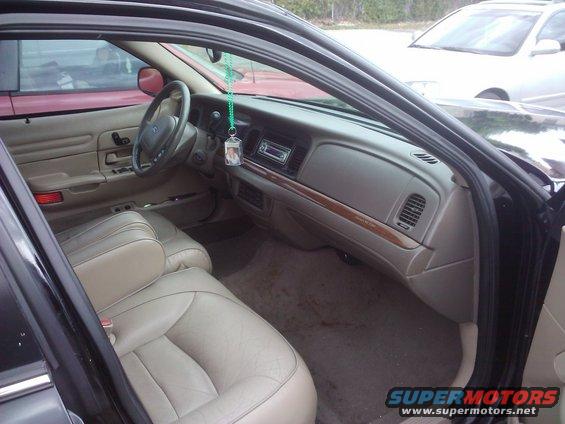 2001 Ford Crown Victoria interior, tan leather seats, dashboard with radio, air vents, and wood trim. Visible in background is a red car and a white car