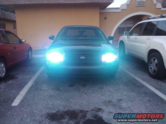 Front view of a black 2001 Ford Crown Victoria with bright headlights on, parked next to a red car and a white SUV