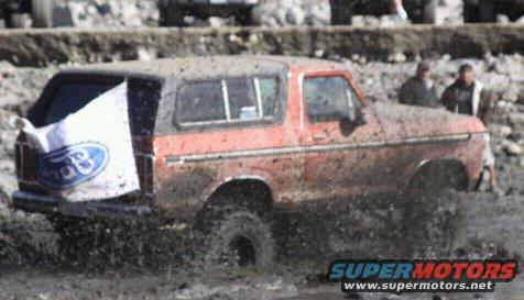 Red 1978 Ford Bronco driving through mud with Ford flag