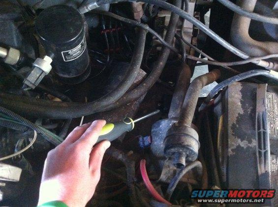 Hand with screwdriver working on engine bay components, including a black cylinder and hoses