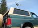 1990 Ford Bronco - A green Ford Bronco with a tan roof trim is viewed from the rear quarter panel, with a helicopter flying in the sky.