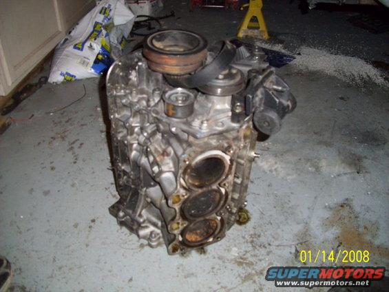 Engine block with cylinders visible, sitting on a concrete floor. A yellow jack stand is in the background