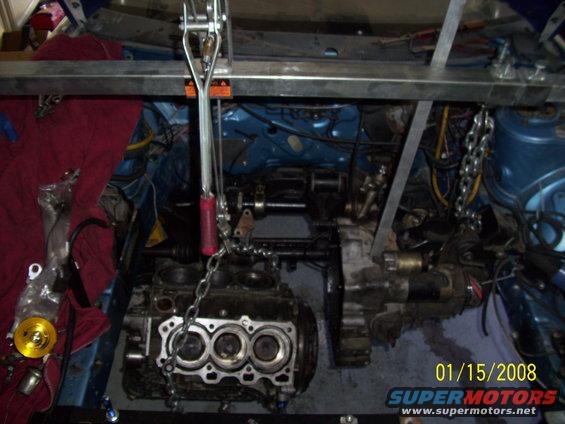 Engine block and transmission assembly suspended by chains and a hoist, with engine bay components visible