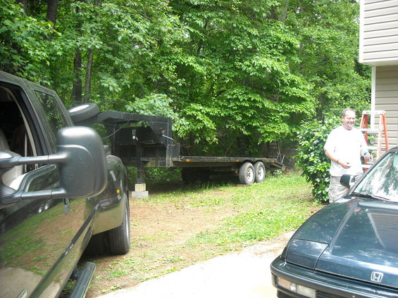 Trailer with dual axles, dark green car in foreground, dark truck in left foreground