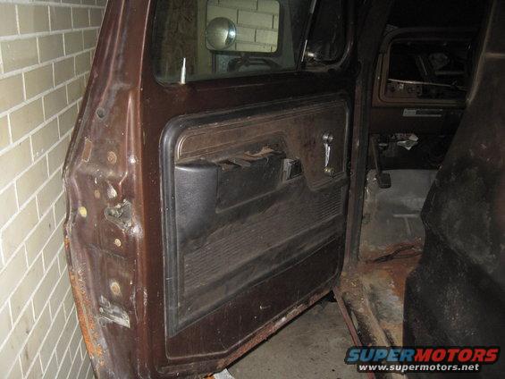 1979 Ford Bronco interior door panel and dashboard view. Shows brown door card, window crank, and part of the dash