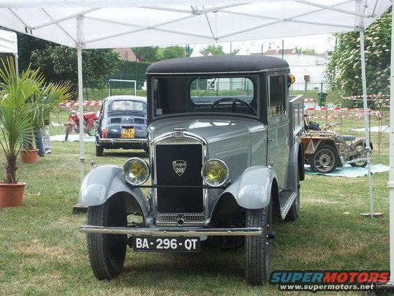 Gray vintage truck with a black roof and bed, parked under a tent. A blue Fiat 500 and a motorcycle are visible in the background