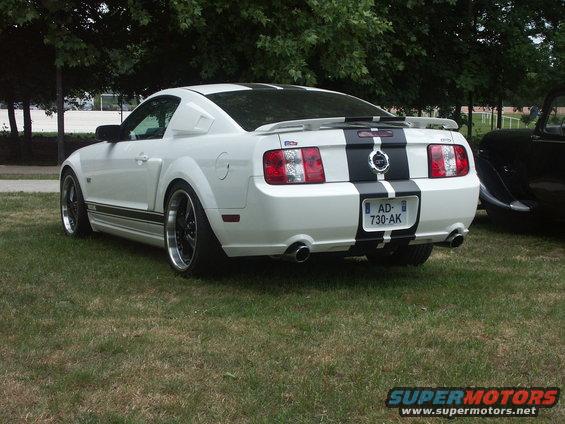 Rear view of a white 2005 Ford Mustang GT with black racing stripes and custom wheels parked on grass. Another black car is partially visible to the right