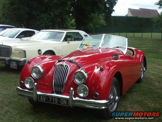 Red convertible sports car with wire wheels and chrome grille parked next to a white Lincoln