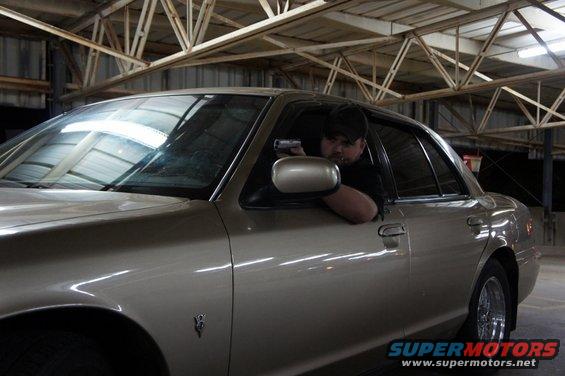 Tan 2000 Mercury Grand Marquis with a man holding a gun in the driver's seat, viewed from the front quarter