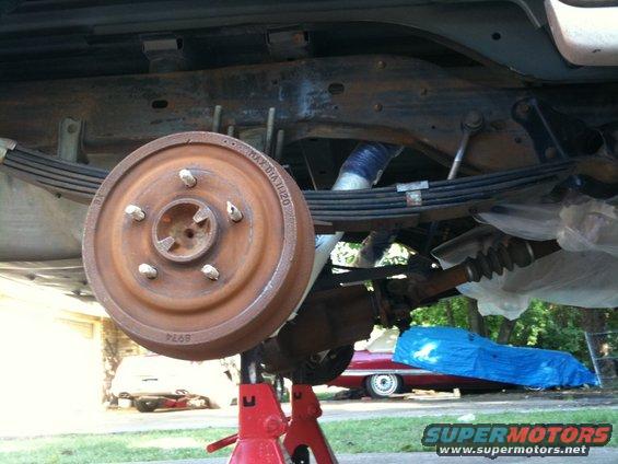 Underside of a vehicle on jack stands, showing the rear axle, leaf springs, and brake drum. Another car is visible in the background