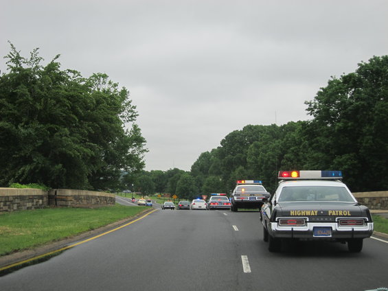 Several highway patrol cars driving in a line on a road with trees on either side