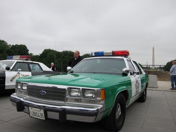 Green police car with light bar, parked next to a black and white police car. Washington Monument in background