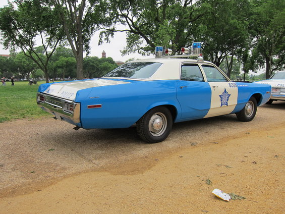 Blue and white police car, rear quarter view, parked on dirt road