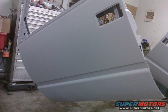 Gray car door with a rectangular cutout, resting on a stand. Another car door is visible to the right