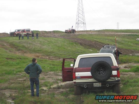 1990 Ford Bronco with spare tire on rear, door open, parked in mud. Other off-road vehicles and people visible on grassy hills