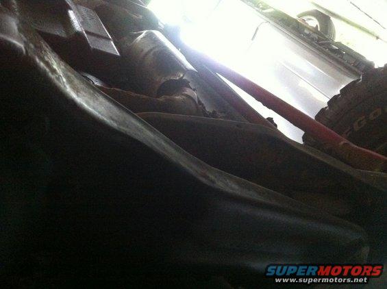 Underside view of vehicle suspension and frame components, with a tire visible in the background