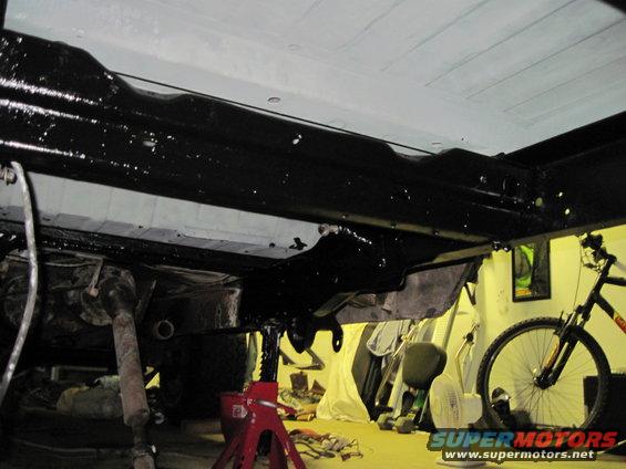 Underside of a vehicle on jack stands, showing black-painted frame and drive shaft. A bicycle is visible to the right