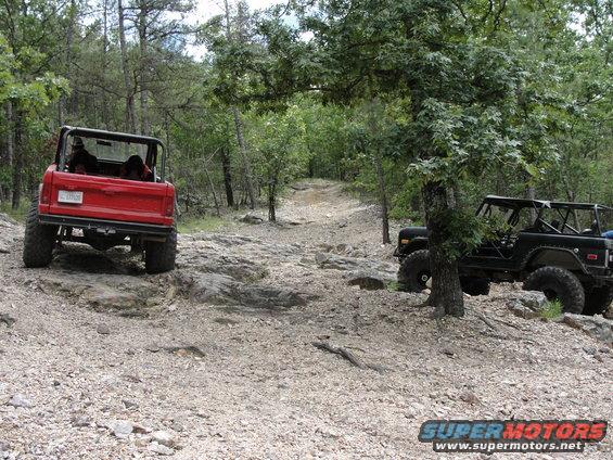 Two off-road vehicles, one red and one black, navigate a rocky, wooded trail