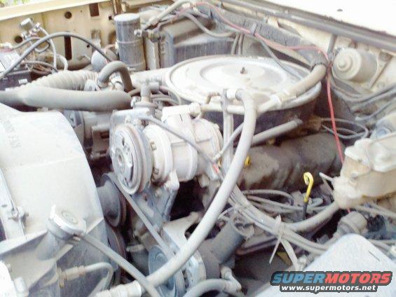 Engine bay of a beige truck, showing the V8 engine, air conditioning compressor, and fan. The engine is dusty and appears to be from the 1980s