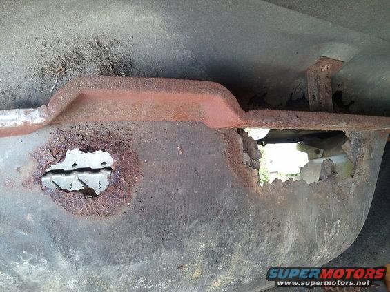 Rusted metal undercarriage with large holes, showing internal components and grass