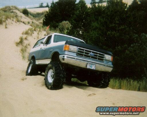 Two-tone blue and white Ford Bronco with lifted suspension and large tires on sand dunes