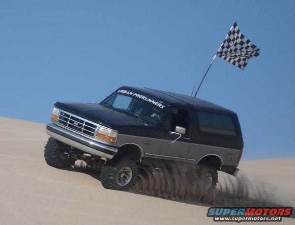 Ford Bronco driving on sand, kicking up dust, with a checkered flag on top