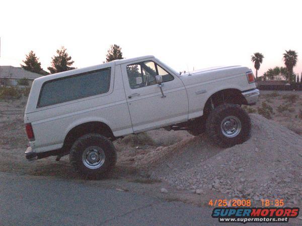 White Ford Bronco with lifted suspension climbing a dirt mound. The vehicle has aftermarket wheels and tires