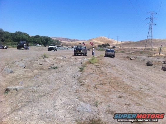 Several off-road vehicles are parked and driving in a dusty, open area with hills and power lines in the background