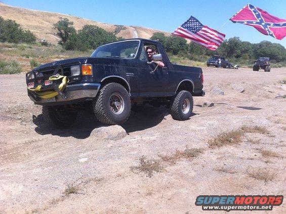 Black 1990 Ford Bronco off-roading over rocks with American and Confederate flags flying