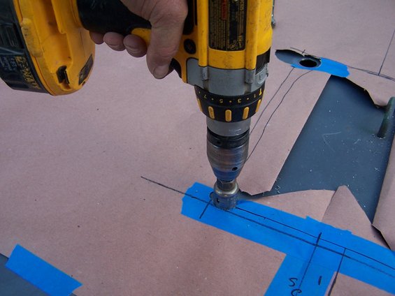 Drill with a hole saw bit cutting into cardboard marked with blue tape and pencil lines